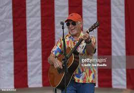Jimmy Buffett stands proudly in front of a flag playing guitar for his fans at a concert.