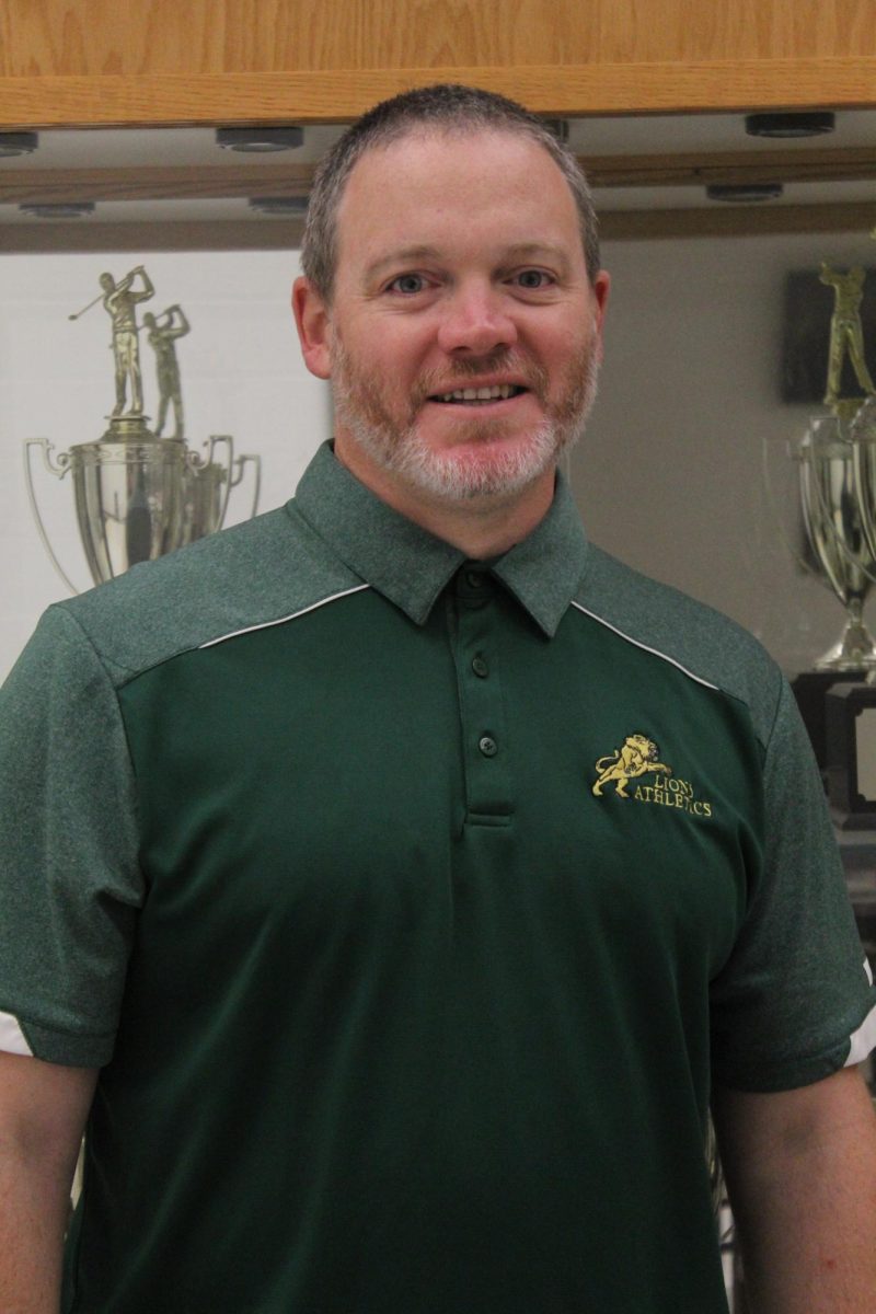 Athletic Director Daniel Barrett smiles in front of the sports trophy case in the cafeteria.

