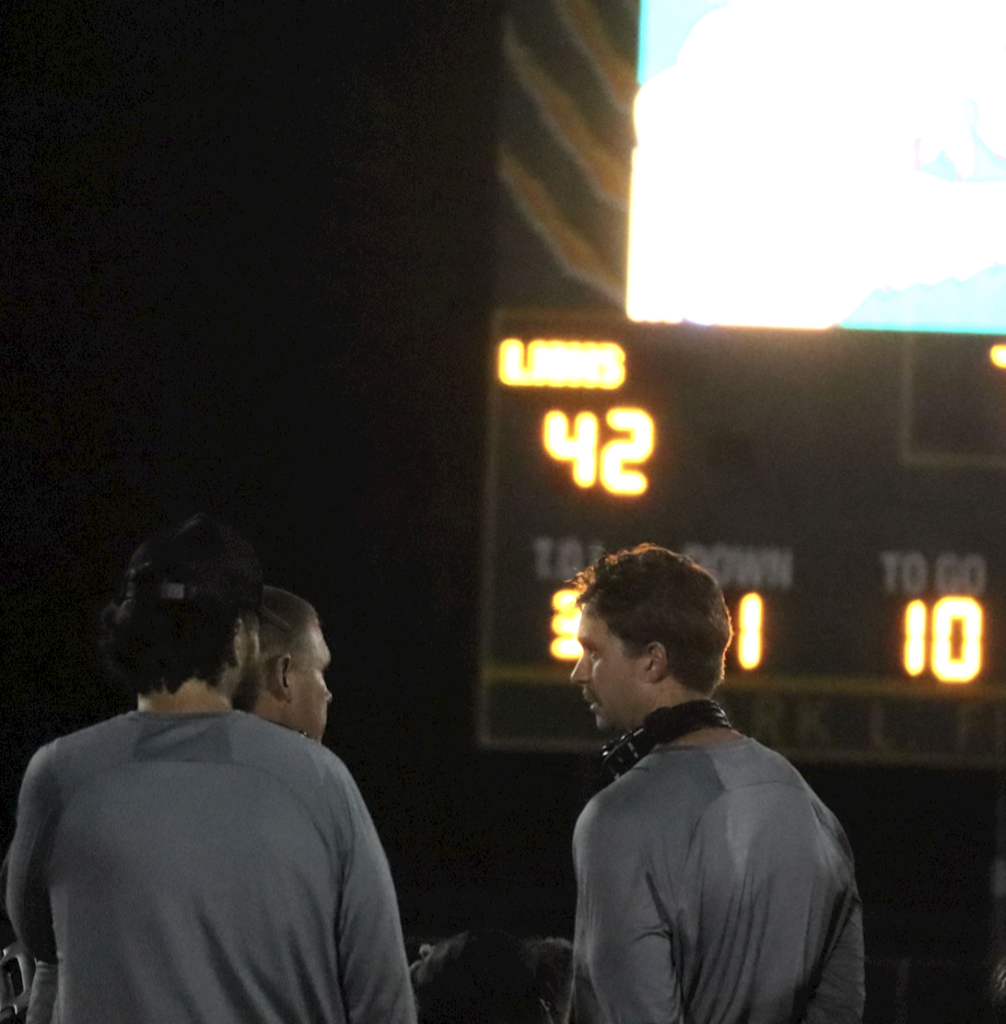 The coaches meet and talk about the game while the score shines in the background.
